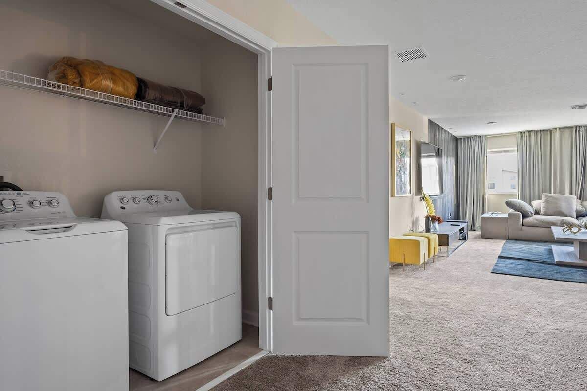 A laundry area with two washing machines, a shelf with a rolled-up towel, and a door leading to a living room with a sofa and a coffee table.