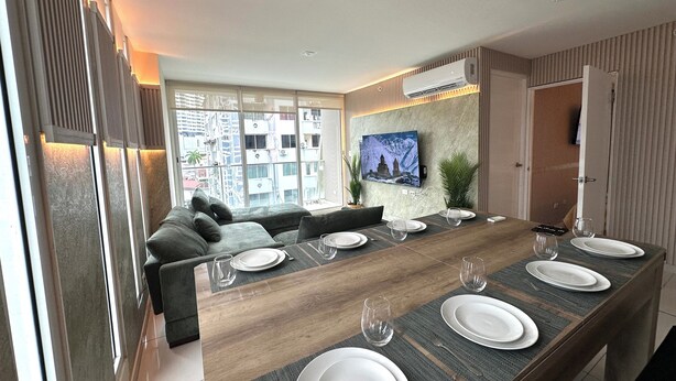 A modern dining area with a long wooden table set for six, a comfortable sofa, and a large flat-screen TV mounted on the wall. - Fabulous 5-bedroom apartment with AC in vibrant Panamá (Panamá)