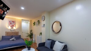 Een kamer met een bed, een slaapbank, een klein tafeltje, een kamerplant, een wandklok en een spiegel.
