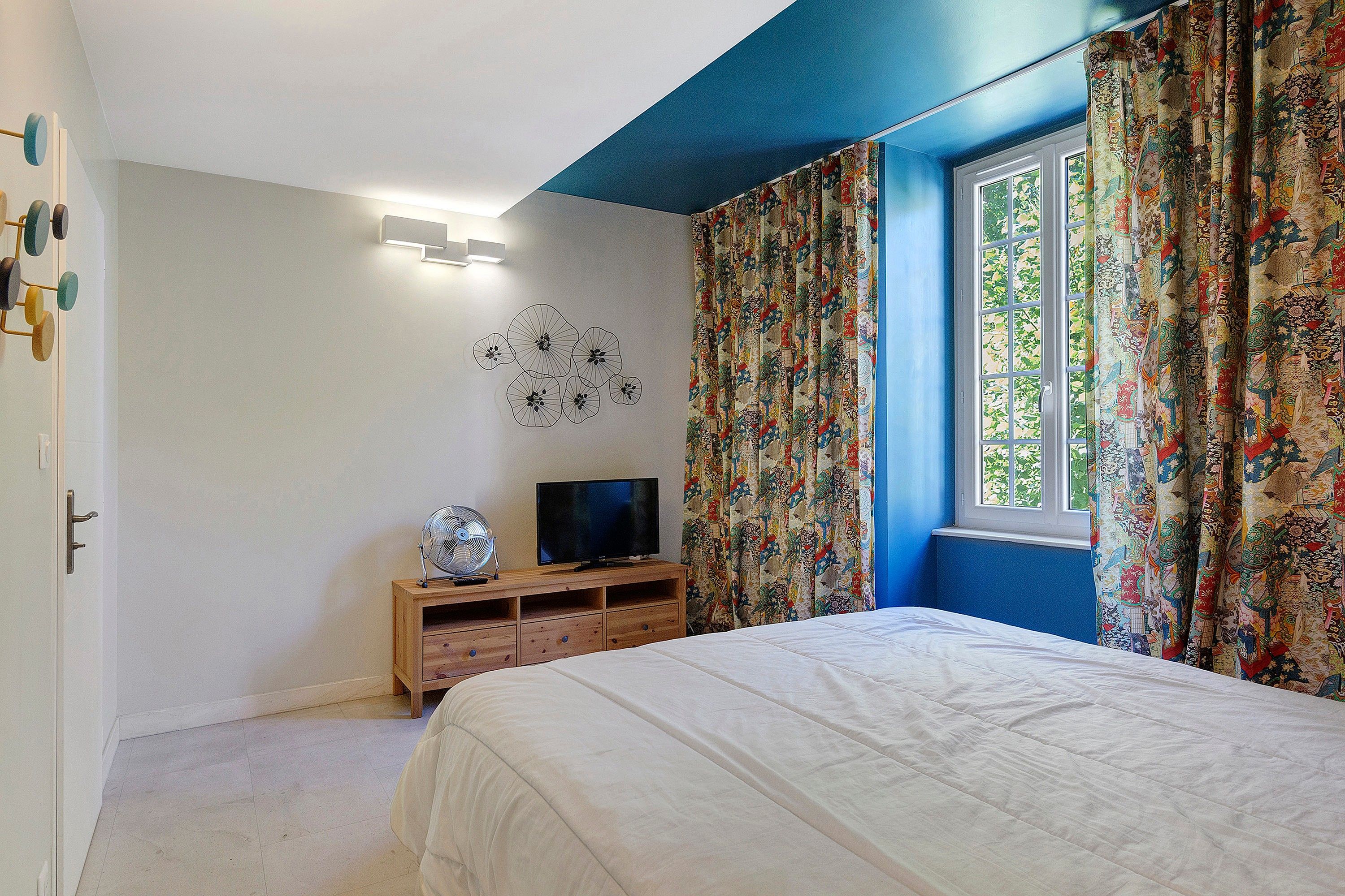 A bedroom with a bed, a TV on a wooden stand, a wall-mounted light, and a window with patterned curtains.