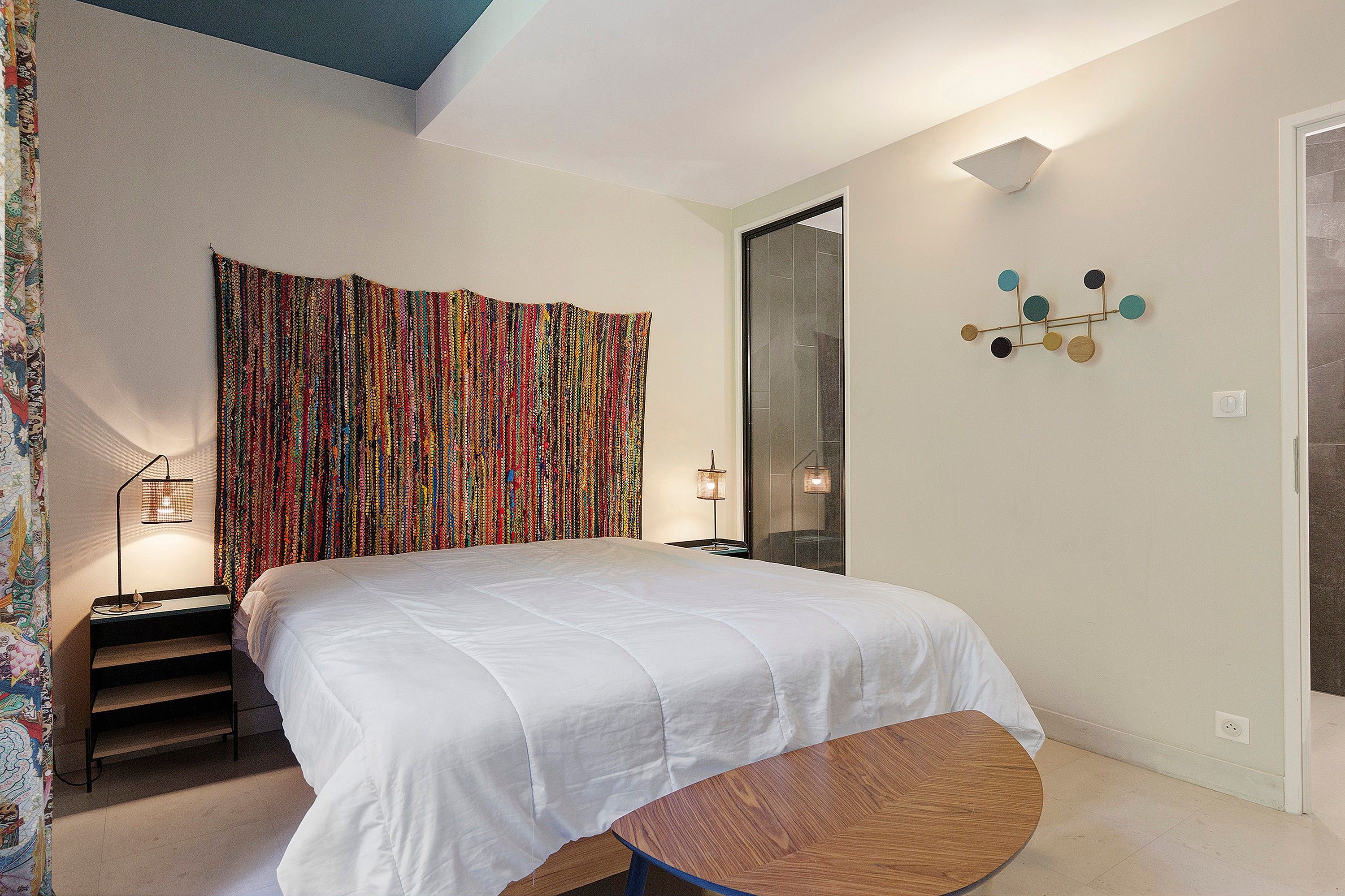 Quarto com uma cama grande, mesa de cabeceira, pequena mesa redonda, cortina colorida e luminária fixada na parede.