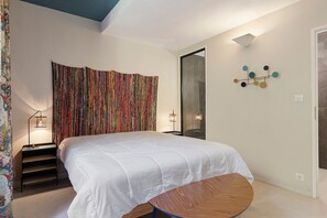 Een slaapkamer met een groot bed, een nachtkastje, een klein rond tafeltje, een kleurrijk gordijn en een wandlamp.