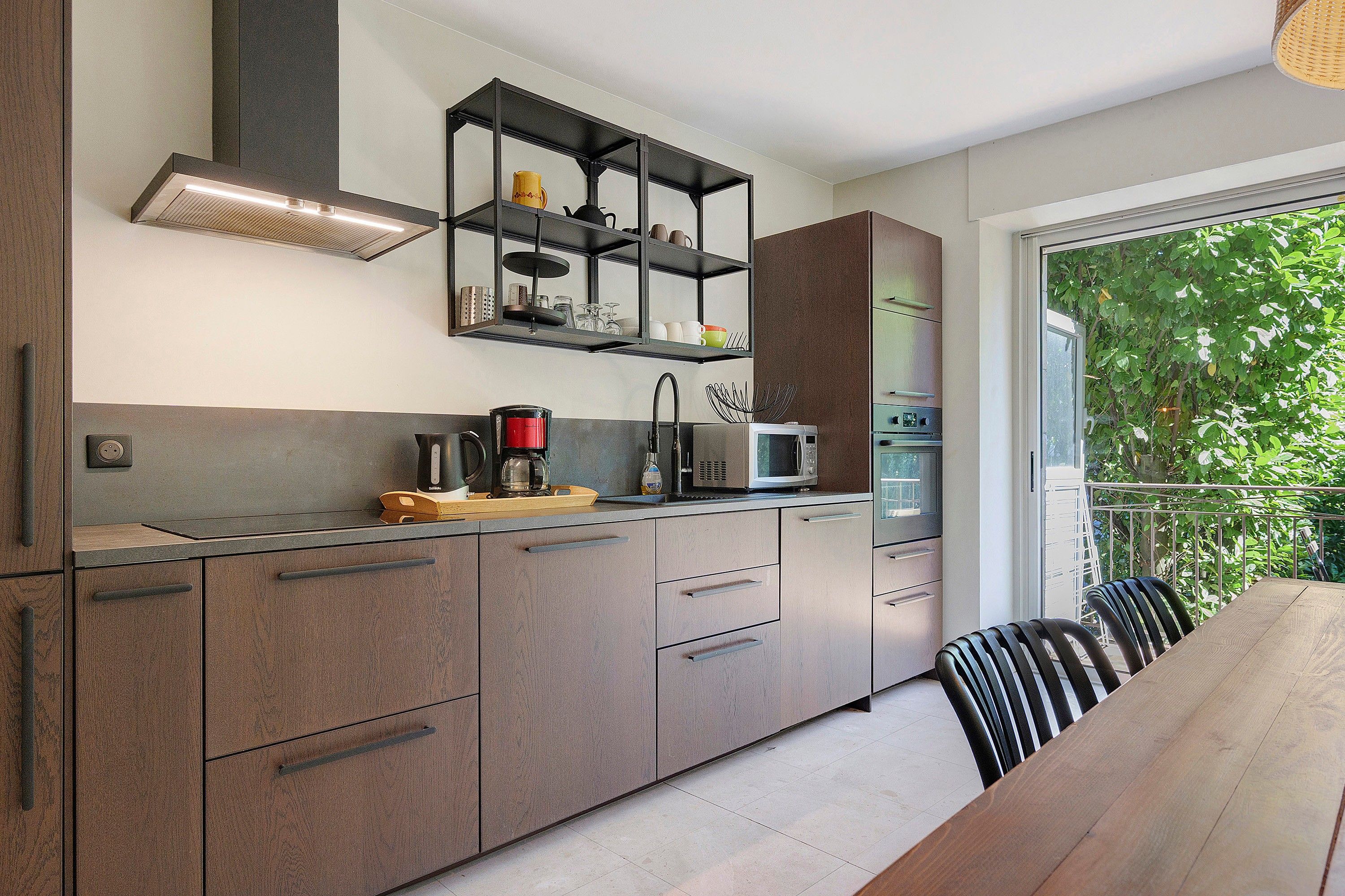Cozinha moderna com eletrodomésticos embutidos, área de jantar com mesa de madeira e vista para o verde através de uma porta de vidro.