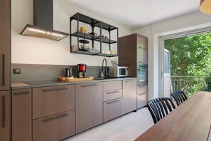 Een moderne keuken met inbouwapparatuur, een eethoek met een houten tafel en uitzicht op groen via een glazen deur.