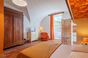 Un dormitorio con cama, armario de madera, televisor sobre un mueble, una silla y una ventana con cortinas.