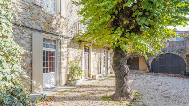 Un edificio in pietra con un cortile sterrato, un albero e un ponte di pietra.