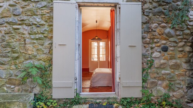 A stone wall with a doorway leading to a room with a bed and a window.