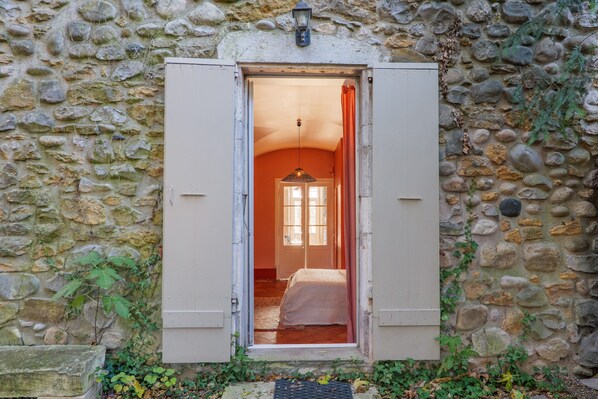Pared de piedra con un vano que da a una habitación con cama y ventana.