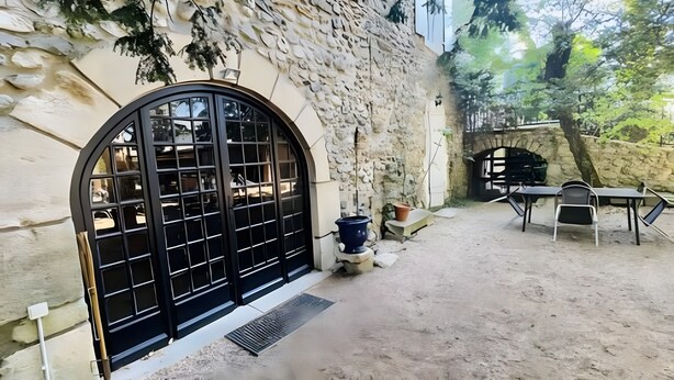 A courtyard with a stone wall, a large arched window, a table with chairs, and a potted plant. - Gîte Yves Apartment with Park View, Private Pool, and Wi-Fi (Vallon-Pont-d'Arc)