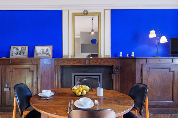 Een eetruimte met een ronde tafel, stoelen, een fruitschaal, een witte beker en een televisie op een standaard.