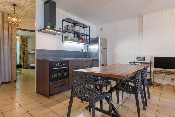 Uma cozinha compacta com forno embutido, micro-ondas e frigorífico. Há uma mesa de jantar com cadeiras e uma TV em móvel.