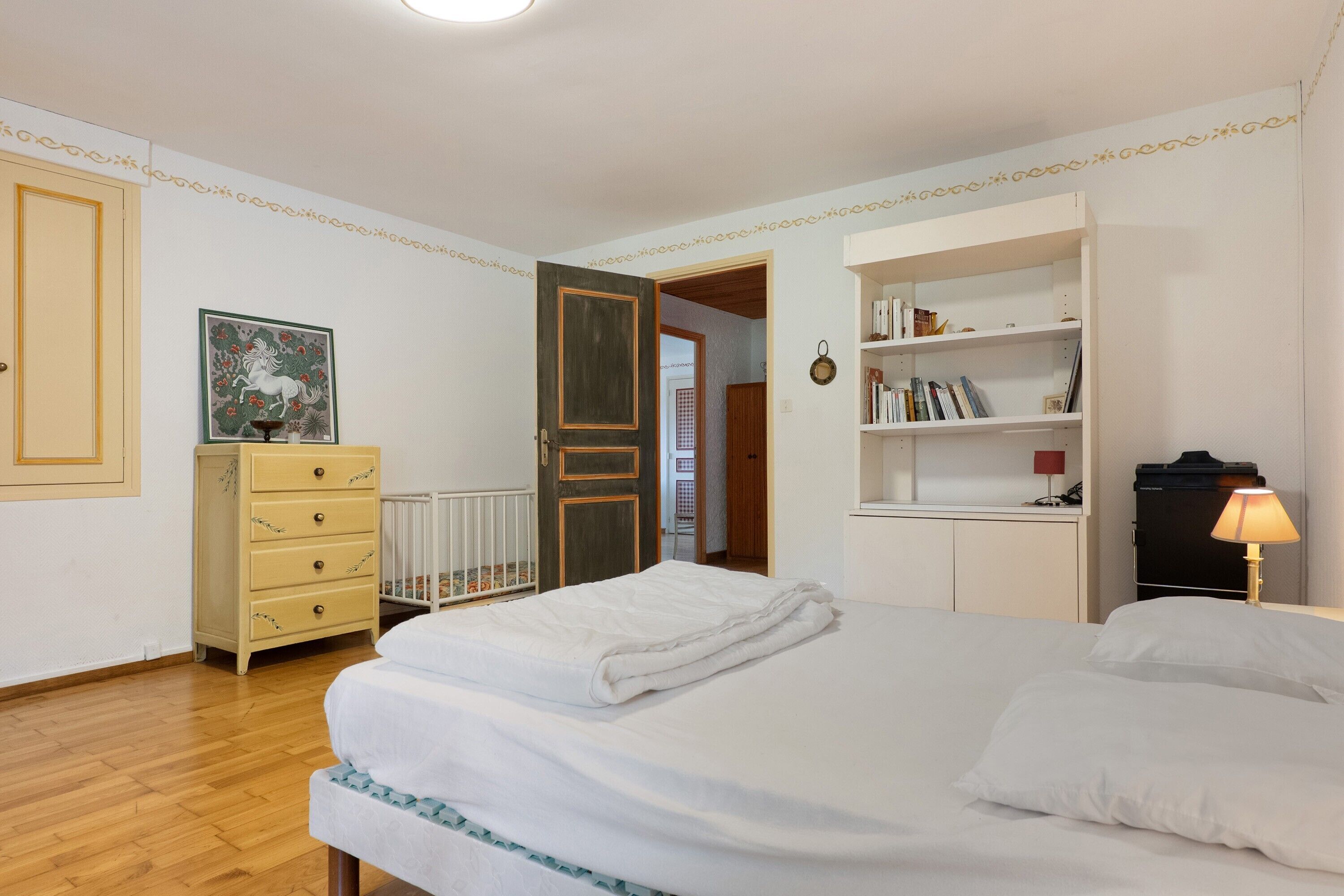 A bedroom with a bed, a dresser, a bookshelf, and a door leading to another room.