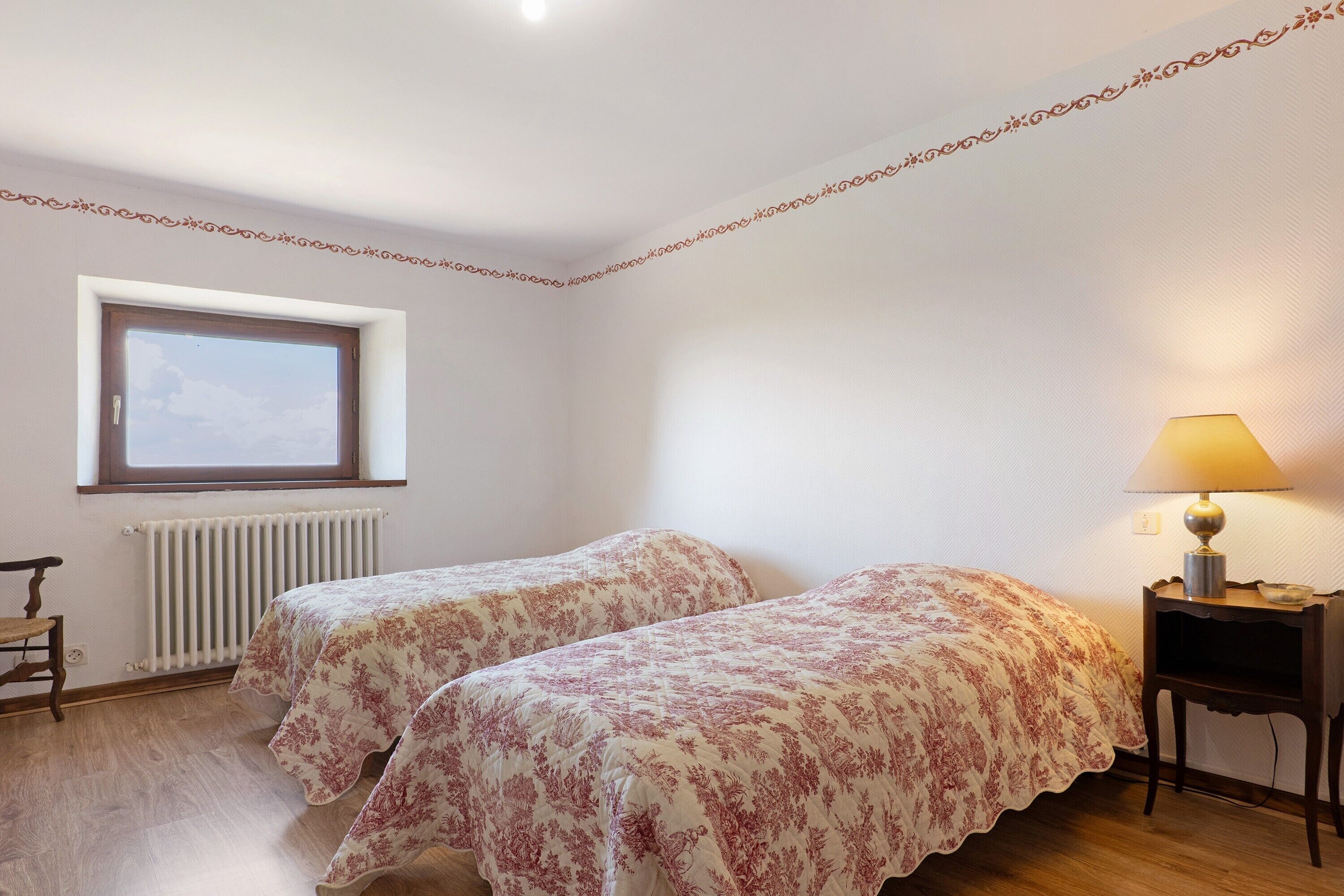 A bedroom with two beds, a window, a radiator, and a bedside table with a lamp.
