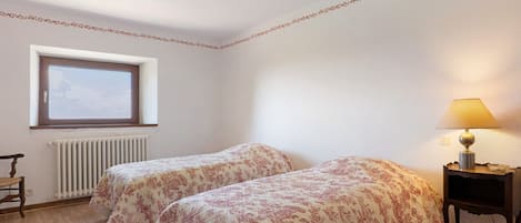 A bedroom with two beds, a window, a radiator, and a bedside table with a lamp.