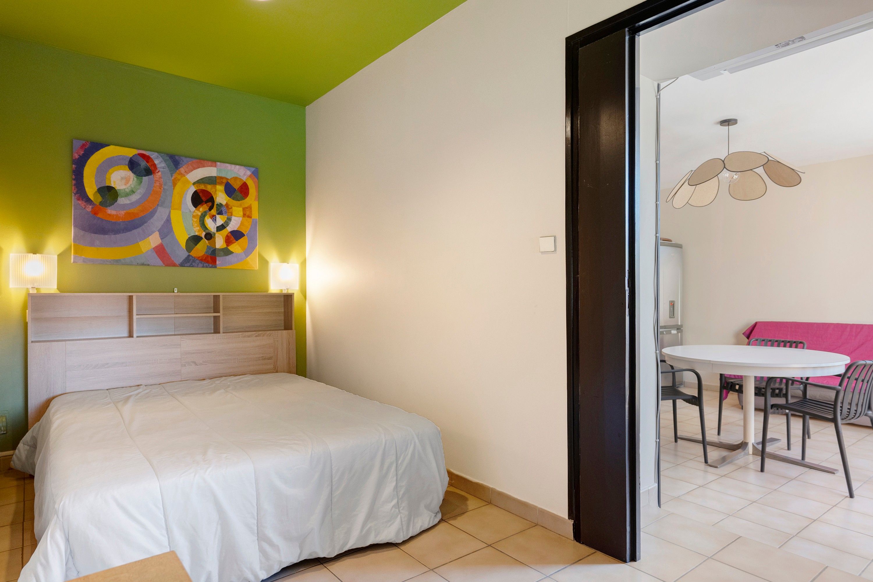 Una camera da letto con un letto, una parete verde, un quadro astratto dai colori vivaci e un'area pranzo con tavolo e sedie.