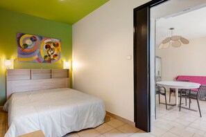 Une chambre à coucher avec un lit, un mur vert, un tableau abstrait aux couleurs vives, et un coin repas avec une table et des chaises.