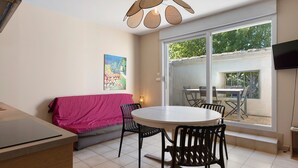 Een zithoek met een ronde tafel en stoelen, een bank met een roze hoes, een schilderij aan de muur en een aan de wand bevestigde televisie.