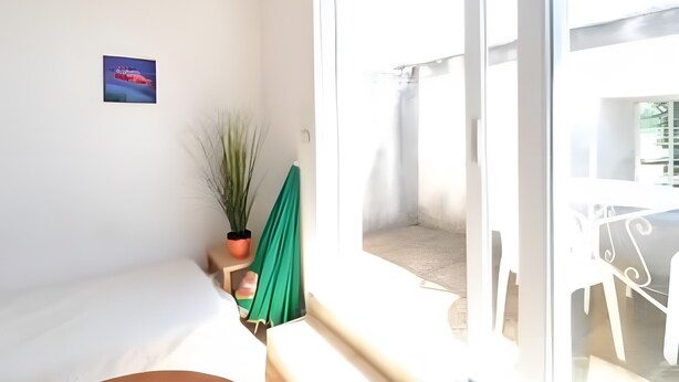 Une pièce comprenant un lit, une fenêtre, une petite table, une plante en pot et un tableau accroché au mur.