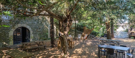 Un patio al aire libre con mesas de picnic y sillas, rodeado de árboles y un muro de piedra.