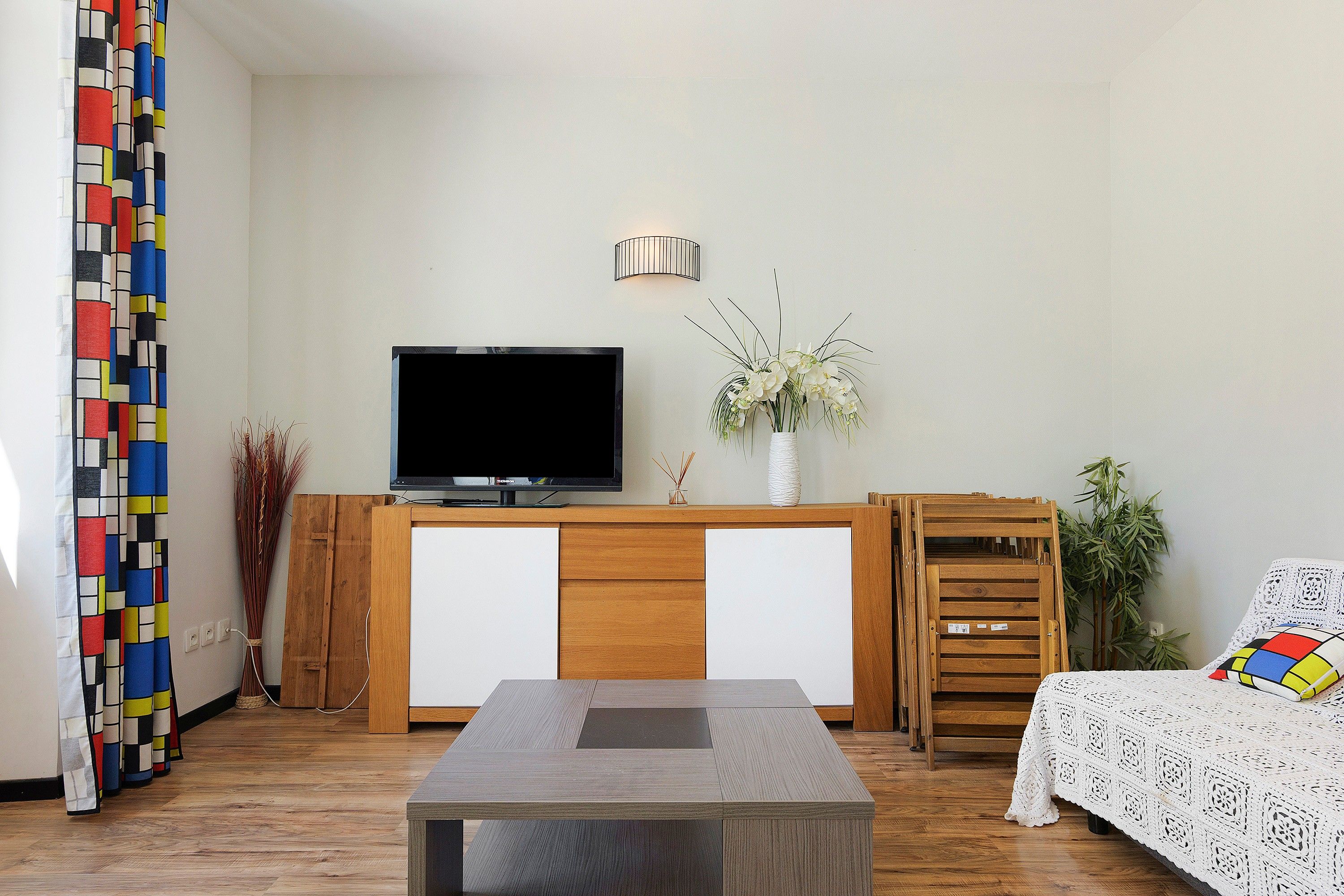 Een woonkamer met een flatscreen televisie, een houten kast, een witte vaas met bloemen en een gestreept bankje.