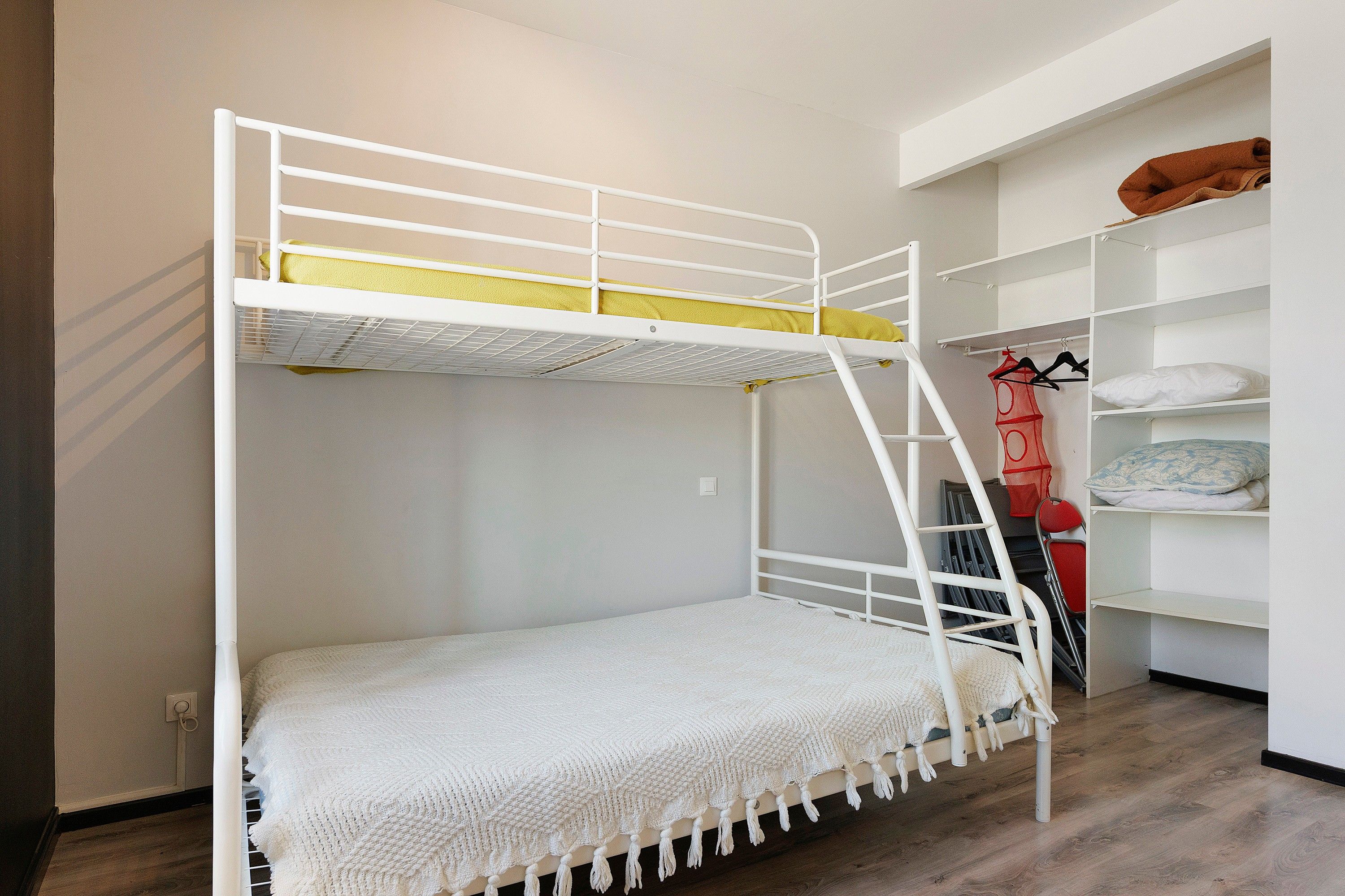 Un lit superposé avec une structure blanche et un matelas jaune. Il y a une couverture blanche sur le lit du bas.