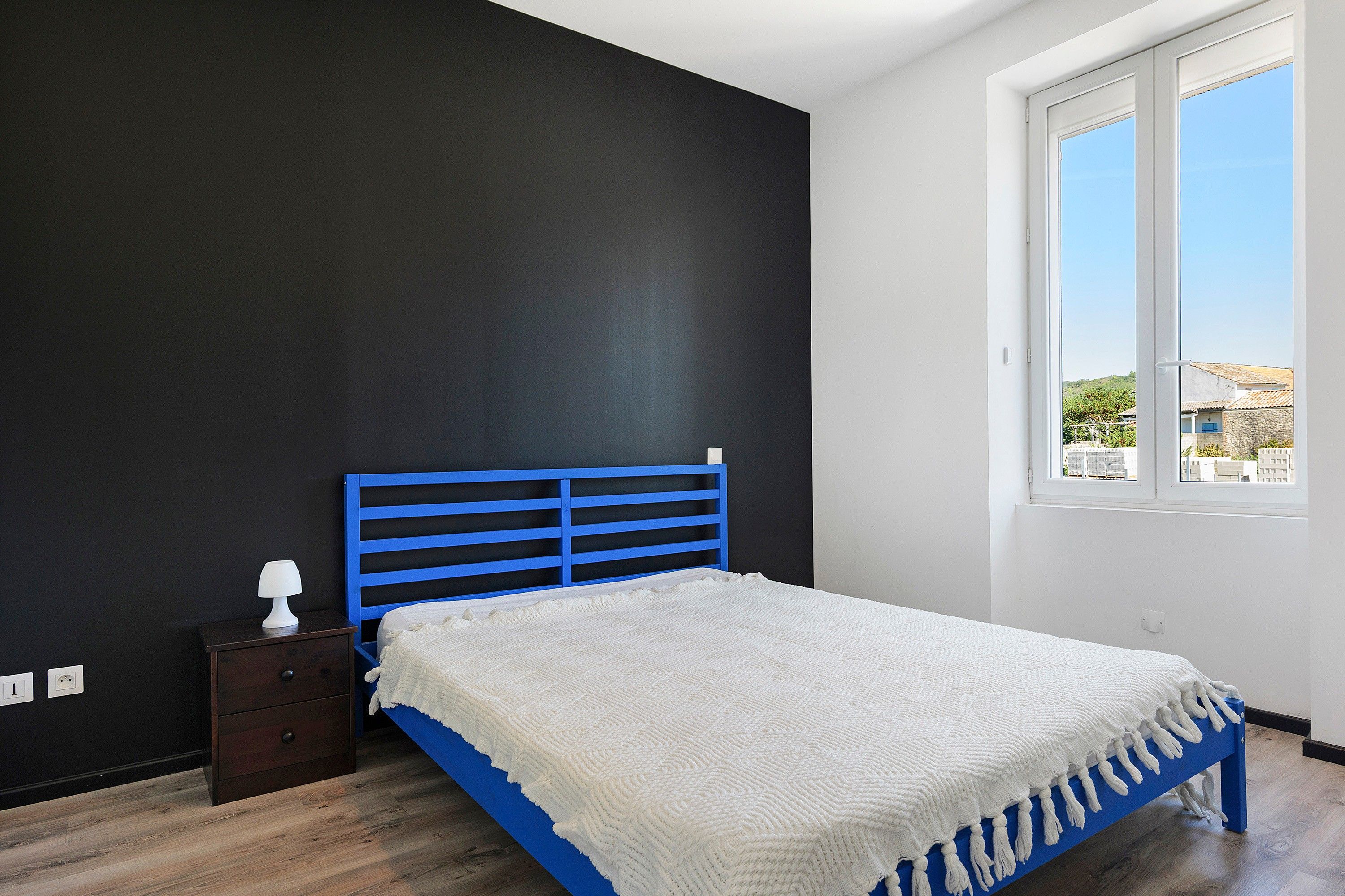 Quarto com estrutura de cama azul, manta branca, mesa de cabeceira com abajur e janela com vista para edifícios.