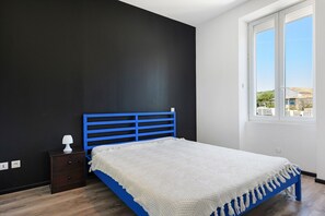 A bedroom with a blue bed frame, a white blanket, a nightstand with a lamp, and a window with a view of buildings.