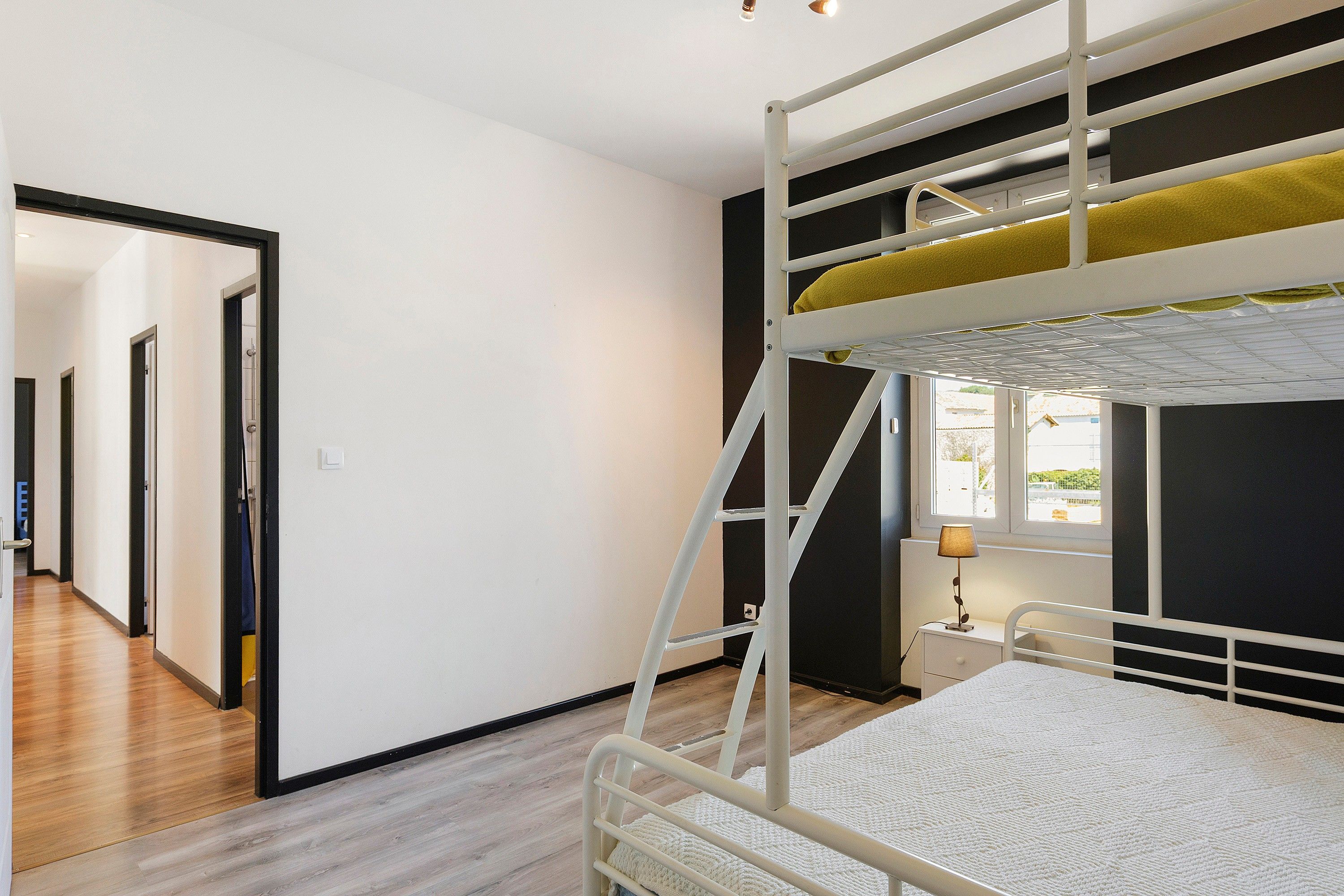 Un lit superposé avec une échelle, une table de chevet avec une lampe et une vue sur l’extérieur par une fenêtre.