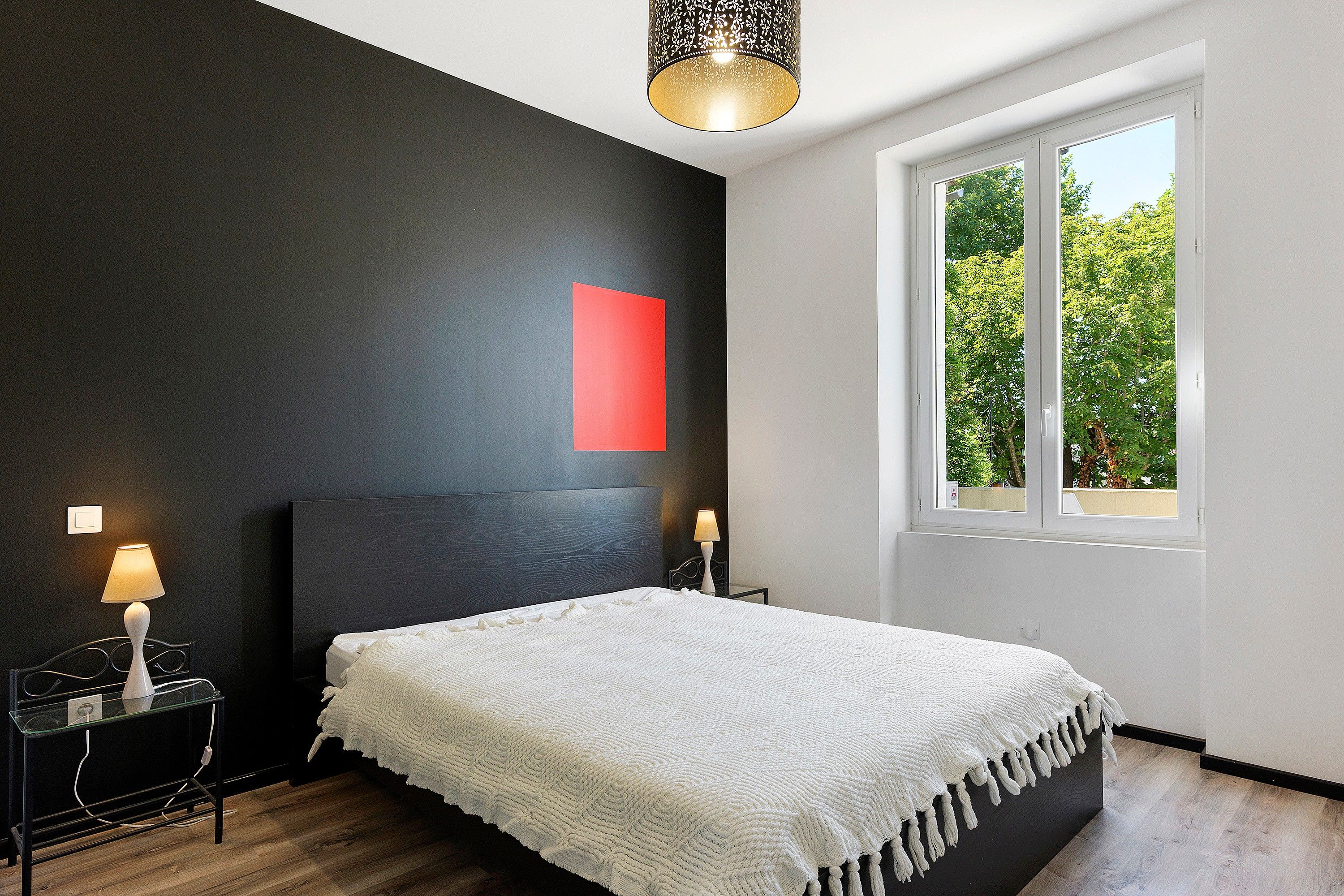 Una camera da letto moderna con un letto grande, due comodini, un tavolino e una finestra che si affaccia su una vista verde, con una parete decorata di rosso.