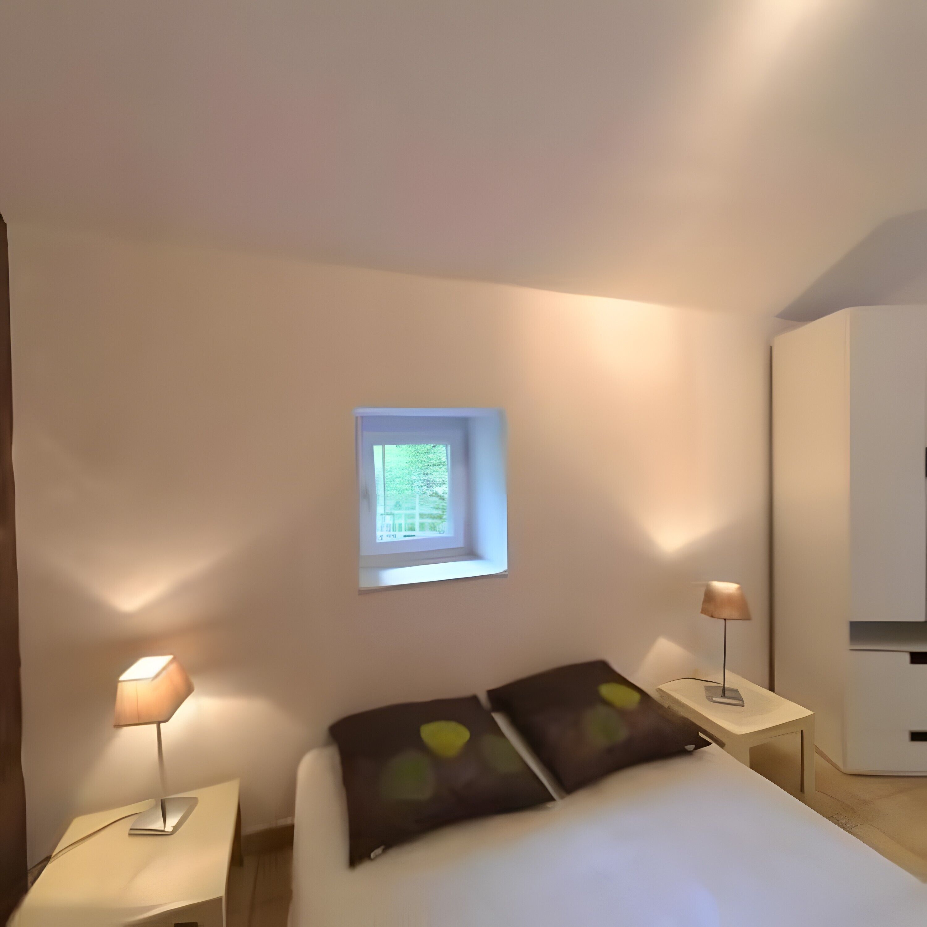 A bedroom with a bed, two bedside lamps, a small window, and a wardrobe.