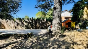 Un área exterior cercada con un árbol, un cobertizo pequeño y un edificio al fondo.