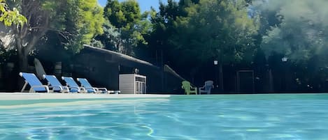 Une piscine à l’eau claire, entourée d’arbres et bordée d’un bâtiment doté d’une zone abritée.