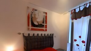 Un dormitorio con cama, mesita de noche, lámpara, un cuadro y una ventana con cortinas.