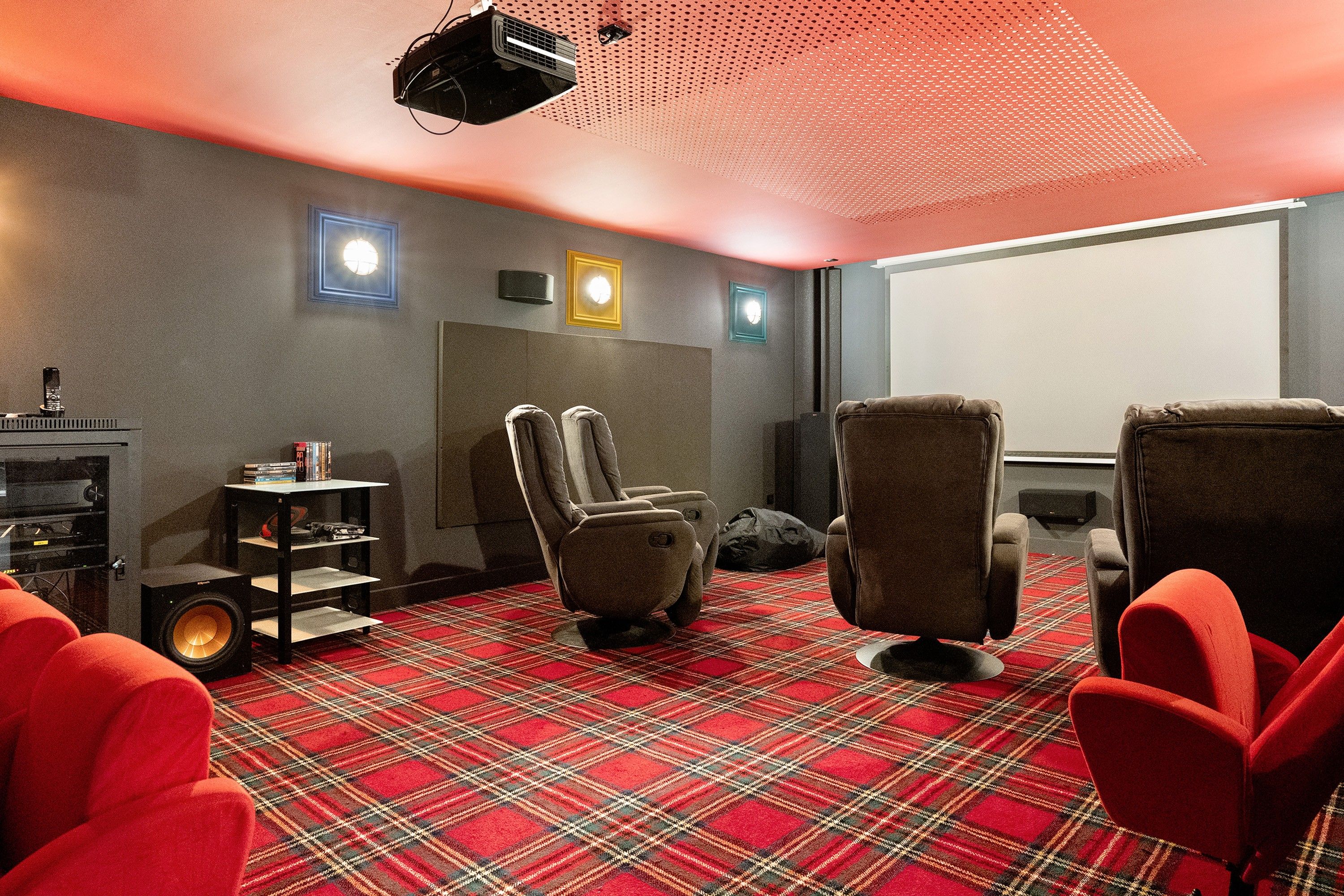 Een kamer met een roodruitig vloerkleed, twee loungestoelen, een klein tafeltje met boeken, een speaker en een aan het plafond gemonteerde beamer.