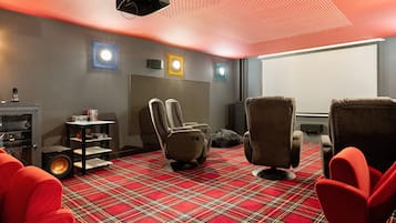 Een kamer met een roodruitig vloerkleed, twee loungestoelen, een klein tafeltje met boeken, een speaker en een aan het plafond gemonteerde beamer.