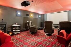 Une pièce avec un tapis à carreaux rouges, deux fauteuils inclinables, une petite table avec des livres, un haut-parleur et un projecteur fixé au plafond.