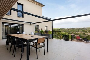 Een overdekt terras buiten met een eettafel en stoelen, een bank en uitzicht op het landschap.