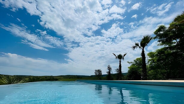 Ein strahlend blauer Himmel mit vereinzelten Wolken, ein Schwimmbecken und üppiges Grün.