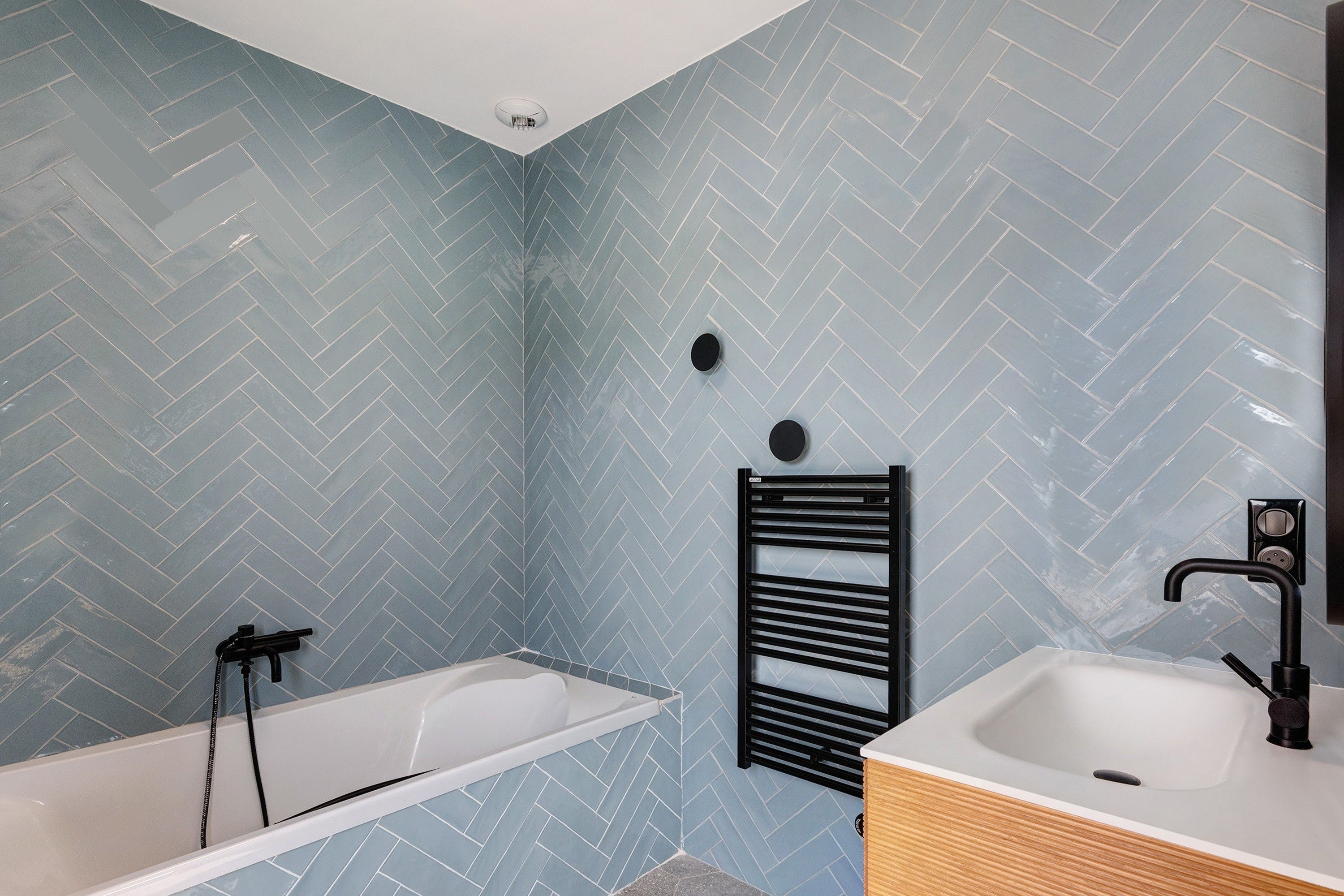 A bathroom with a herringbone-patterned tile wall, a black towel rack, a white bathtub, and a wooden vanity with a sink.