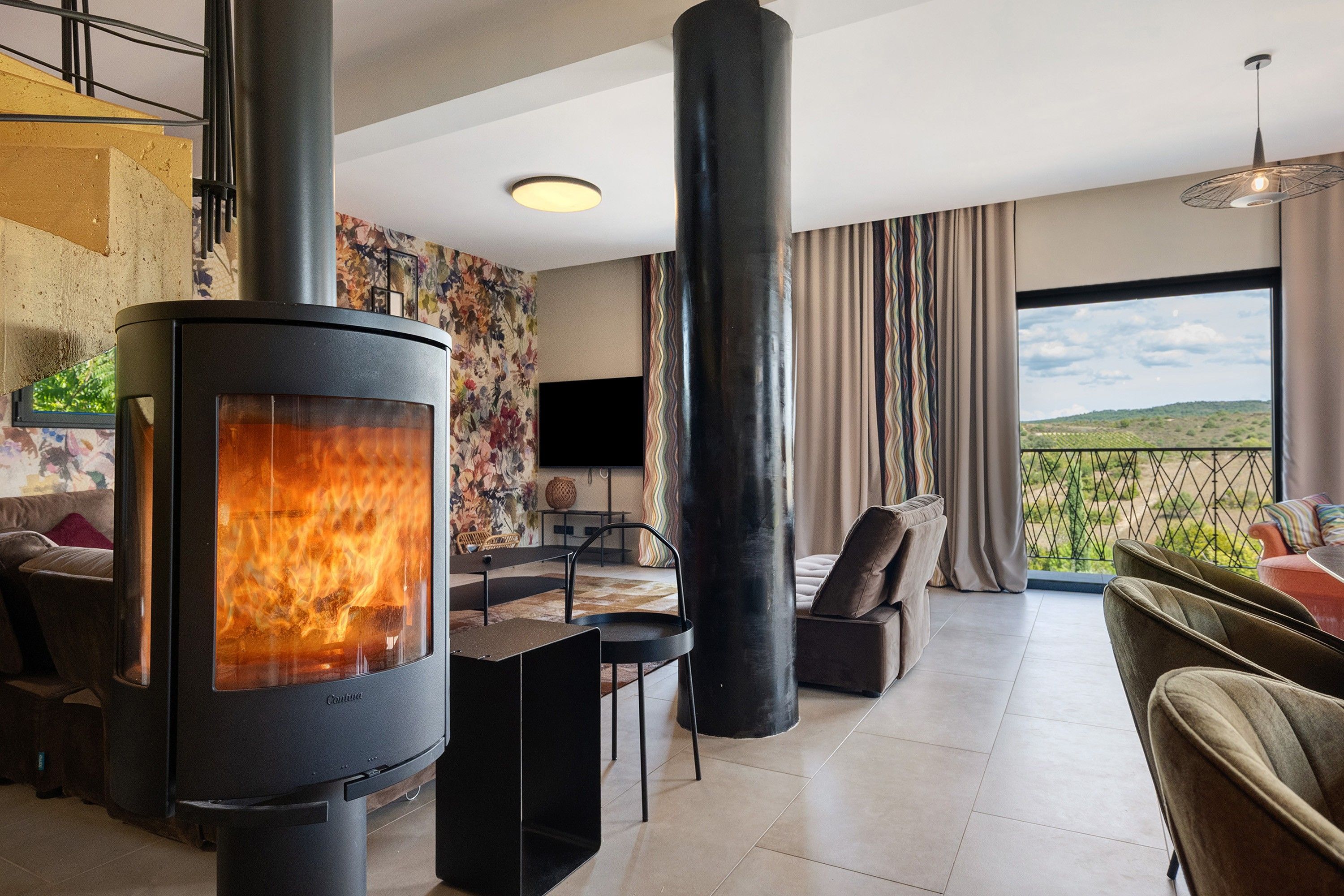 A modern living room with a wood-burning stove, a flat-screen TV, and a view of the countryside.