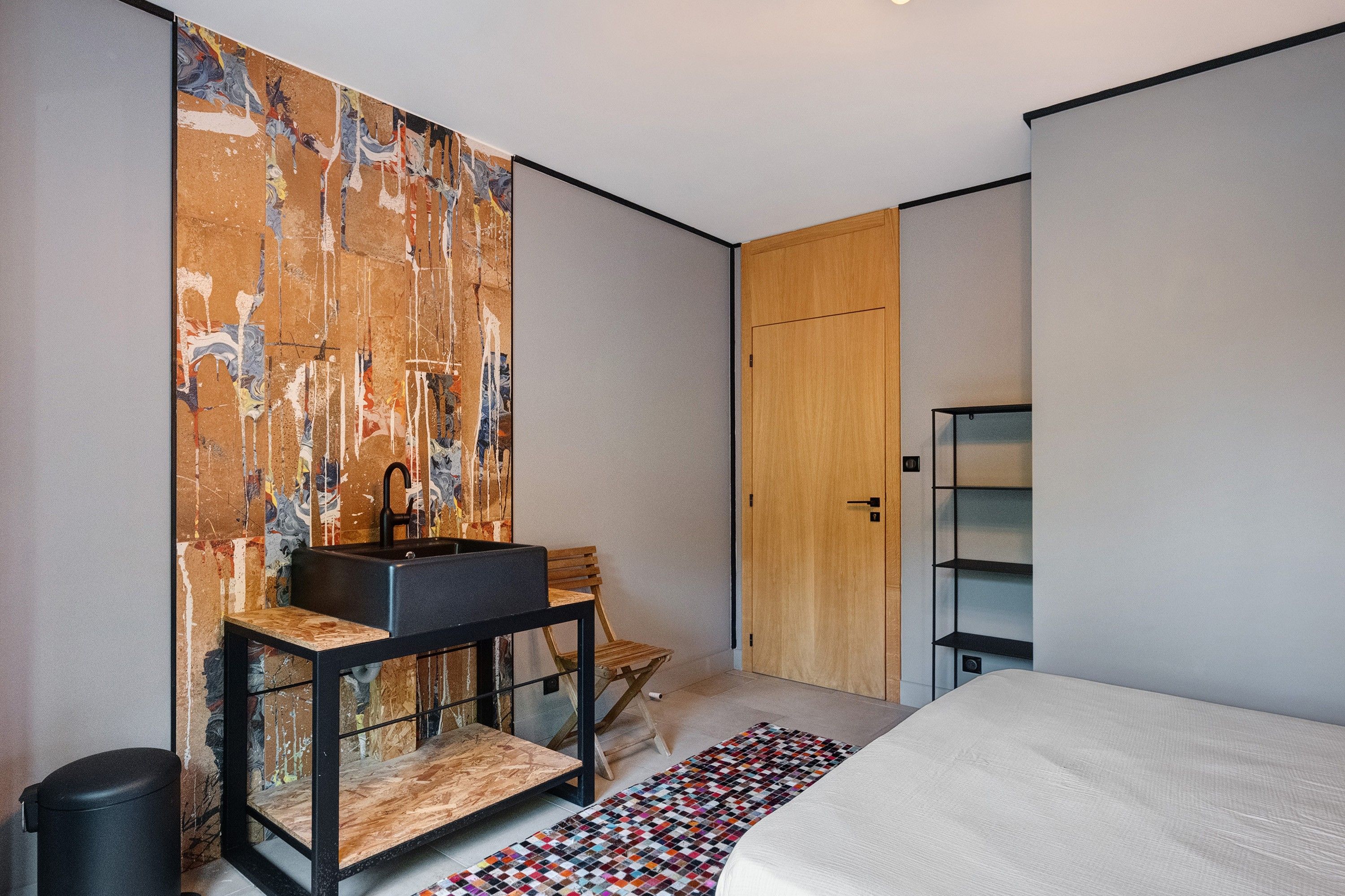 Een moderne slaapkamer met een groot bed, een houten garderobe, een zwarte wastafel op een houten tafeltje en een kleurrijk vloerkleed.