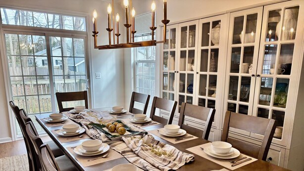 Un comedor con una mesa larga dispuesta para seis personas, rodeada de sillas, con un candelabro encima y un aparador con puertas de cristal al fondo.