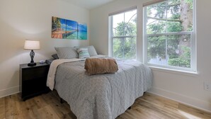 Une chambre avec une grande fenêtre, un lit recouvert d’une couvre-lit à motifs blancs et gris, une table de chevet avec une lampe et un tableau accroché au mur.