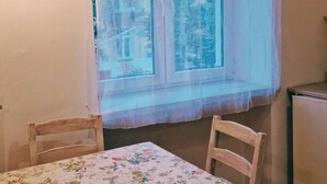 Ein Zimmer mit Esstisch, zwei Stühlen, einem Fenster mit Blick auf Gebäude und einer Wandleuchte.