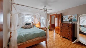 Una cama con dosel sostenido por cuatro columnas, con estructura de madera y una colcha verde. A la derecha, un aparador de madera con varios cajones y un espejo. Un cuadro con motivos florales decora la pared sobre el aparador.