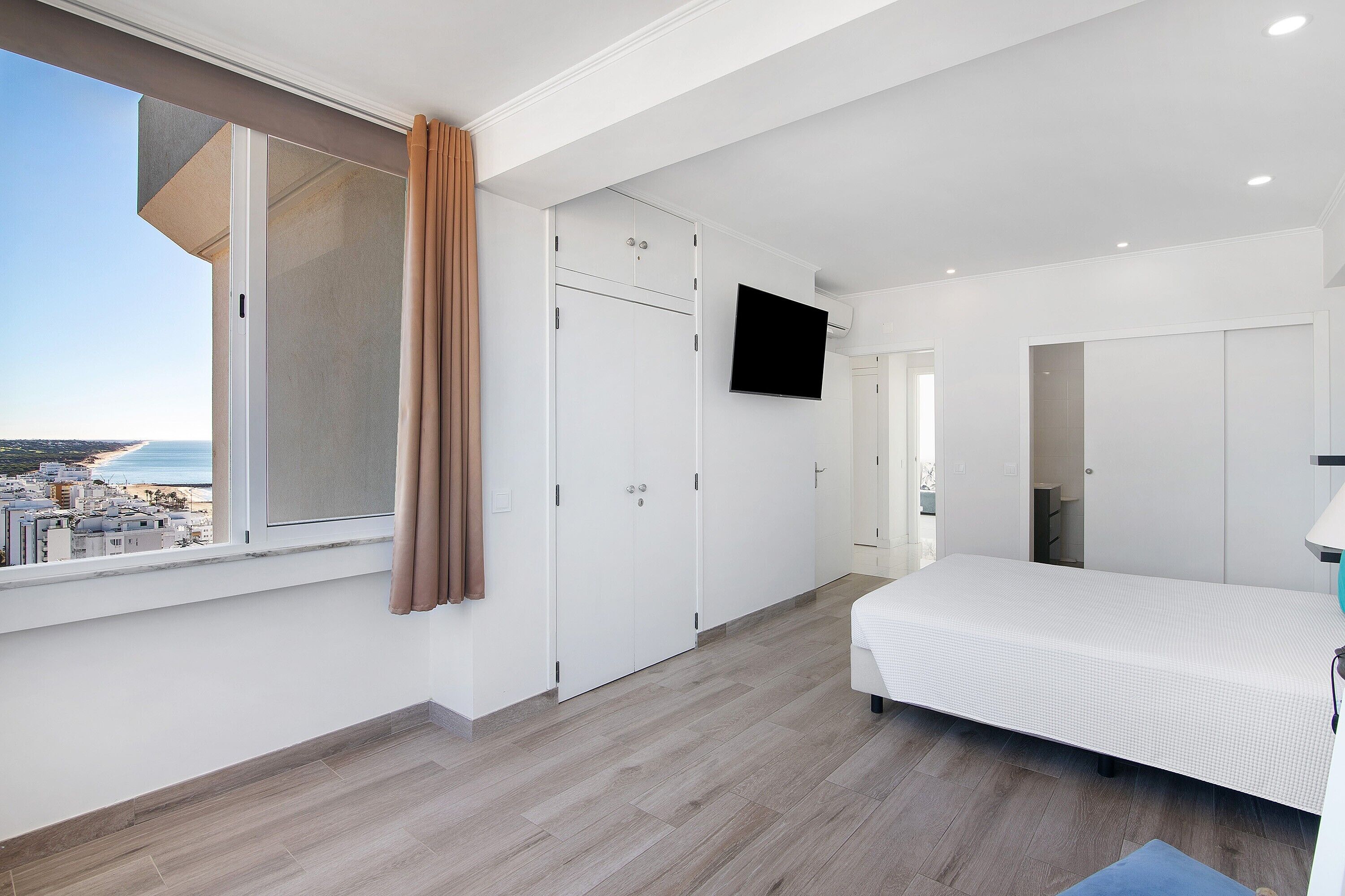 Ein Schlafzimmer mit Bett, Kleiderschrank, Fernseher und einem Fenster mit Blick auf Gebäude.