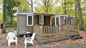Une petite cabane avec une terrasse en bois et deux chaises blanches à l’extérieur.