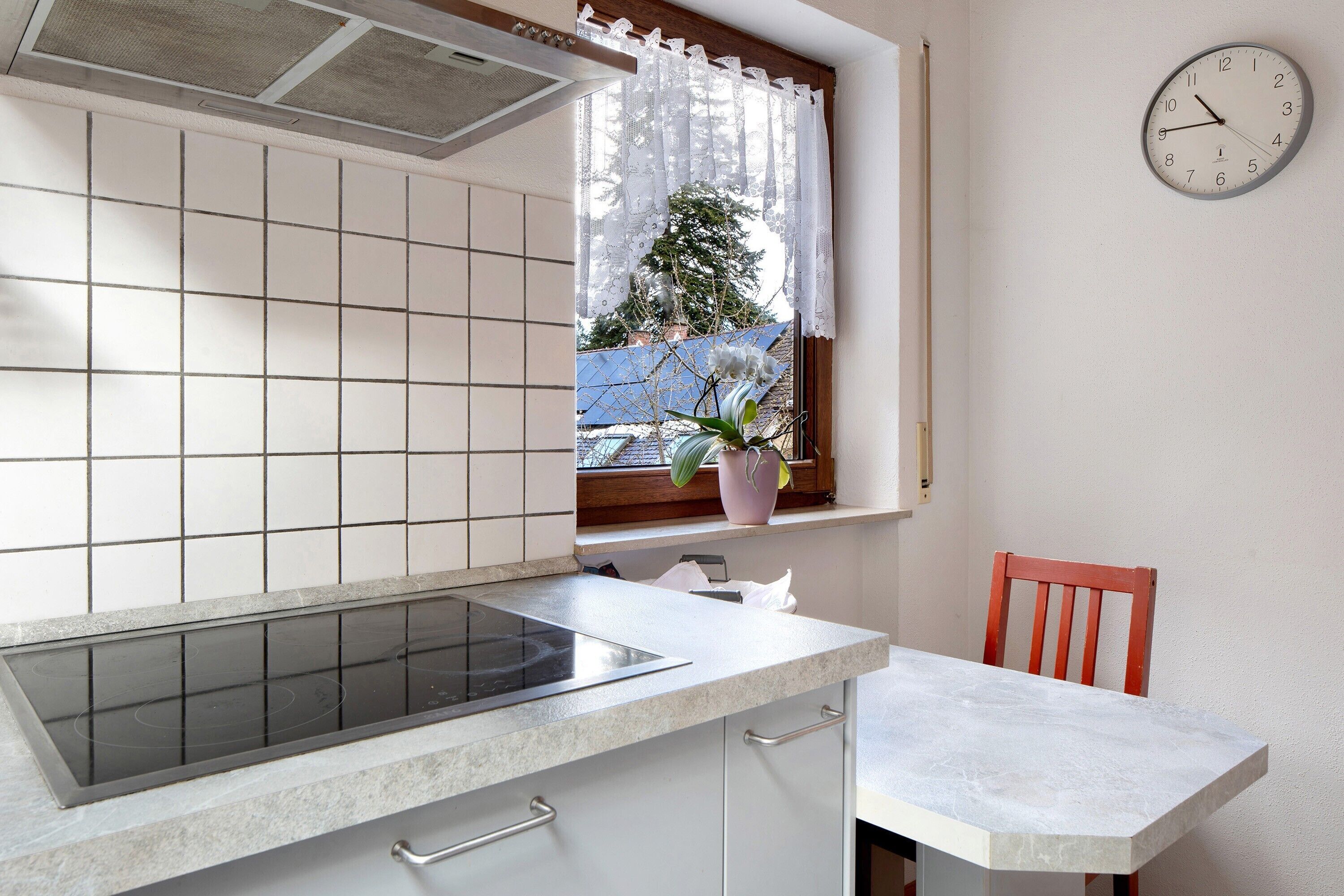 A kitchen with a stove, a window with a curtain, a clock, and a chair.