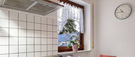 A kitchen with a stove, a window with a curtain, a clock, and a chair.