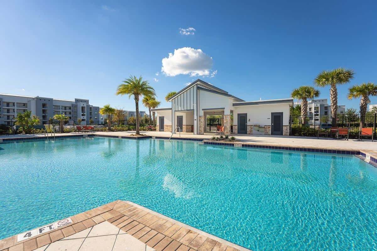 En swimmingpool med en djupmarkering som visar 1,2 meters djup, omgiven av palmer och moderna byggnader.